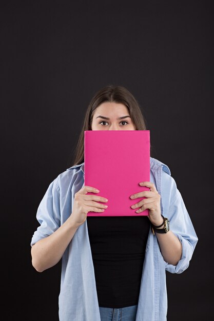 Foto hermosa estudiante en camisa vaquera con cabello largo, en la pared negra, asomándose por detrás del libro