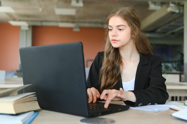 Una hermosa estudiante caucásica está estudiando en la universidad de forma remota Está sentada con una computadora portátil y un bloc de notas y concentrada está viendo una lección de videoconferencia