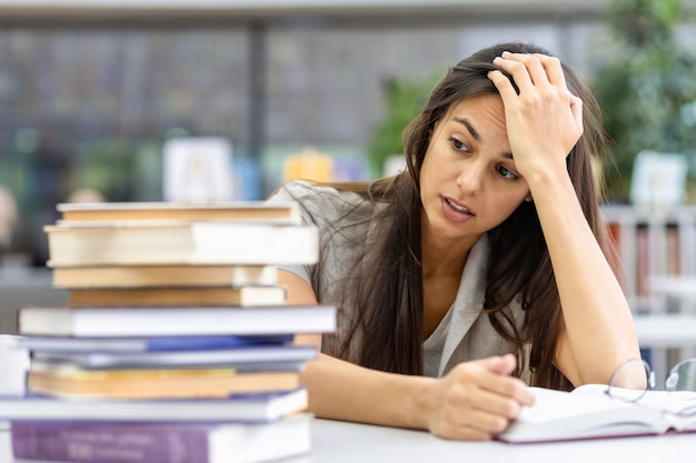 Foto hermosa estudiante caucásica resentida por la gran cantidad de material educativo sentado en la biblioteca