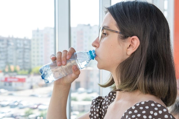 Una hermosa estudiante con gafas bebe agua de una botella de plástico mirando por la ventana Un día caluroso y soleado Reponer la humedad en el cuerpo con agua