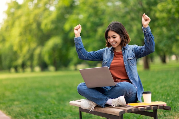Hermosa estudiante de Oriente Medio celebrando el éxito con una laptop al aire libre