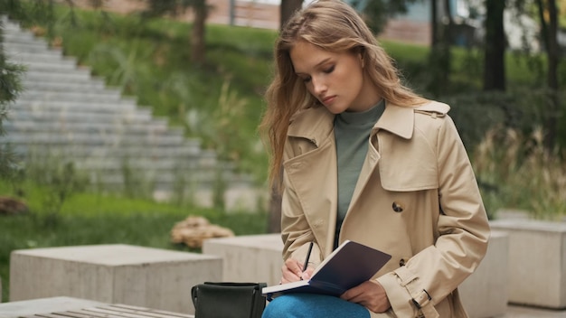 Hermosa estudiante que parece concentrada haciendo los deberes para la universidad en un banco en el parque de la ciudad. Elegante chica rubia estudiando al aire libre