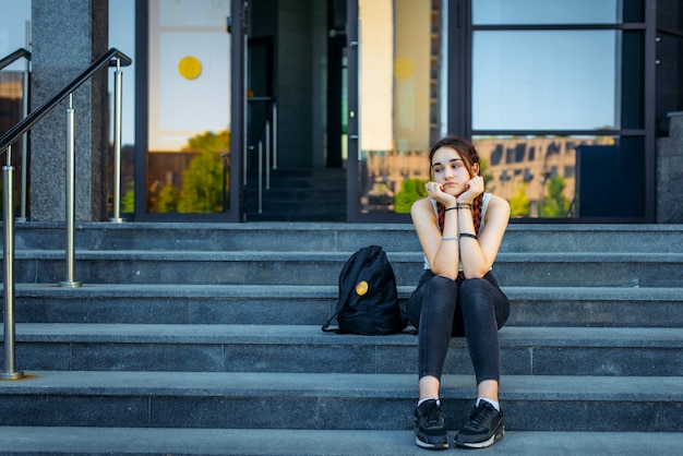 Hermosa estudiante reflexivo sentado en las escaleras al aire libre entre clases. La vida estudiantil, el concepto de educación.