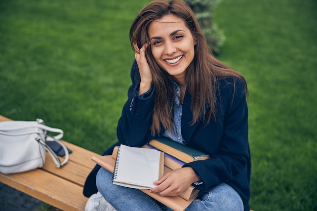 Hermosa estudiante sentada en un banco con el estudio de materiales de rodillas y sonriendo