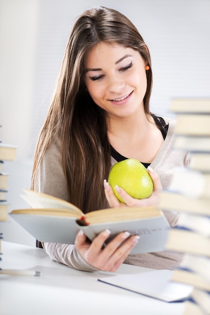 Hermosa estudiante sentada con manzana entre muchos libros y aprendiendo.