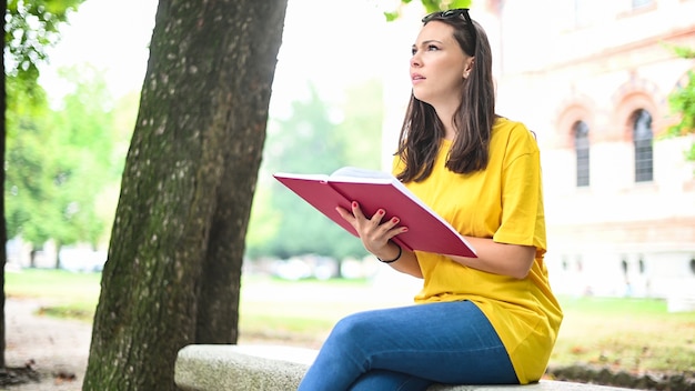 Hermosa estudiante universitaria leyendo un libro en un banco en un parque