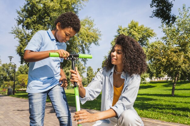 Hermosa familia afroamericana feliz uniéndose en el parque - Familia negra divirtiéndose al aire libre, mamá enseñando a su hijo a montar en scooter