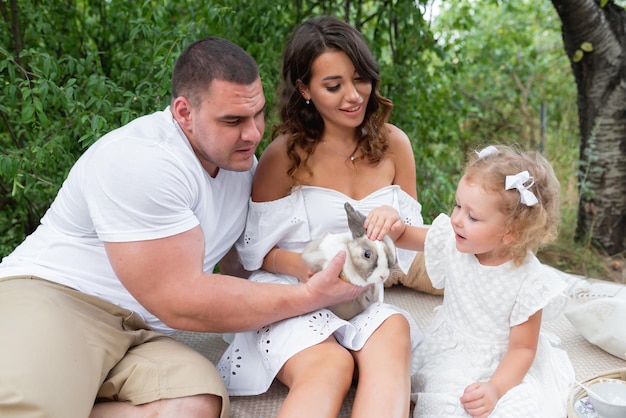 Hermosa familia amorosa divirtiéndose al aire libre Mamá, papá y su pequeña hija están sentados en la naturaleza sosteniendo un conejo blanco en sus manos