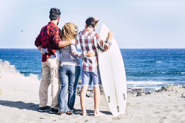 Hermosa familia junta para siempre en la playa con un día soleado y adolescente surfista - mirando el mar y el cielo con mesa de surf y gente feliz - vacaciones de verano - vista trasera