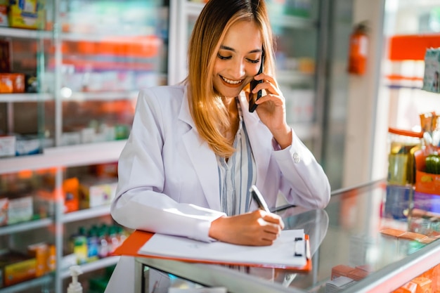 Foto una hermosa farmacéutica haciendo una llamada mientras usa un bolígrafo escribiendo una solicitud de nombre de medicamento en papel