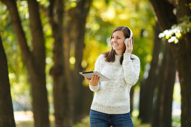 La hermosa feliz alegre mujer de cabello castaño en suéter blanco con una tableta escuchando música en los auriculares blancos en el parque de otoño en un día caluroso. Otoño en la ciudad.