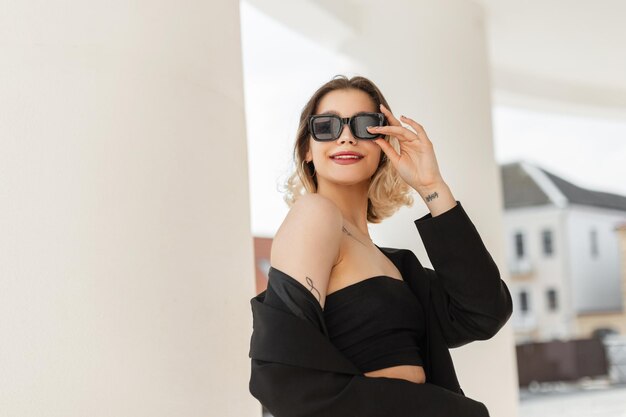 Hermosa feliz elegante joven modelo de mujer elegante con una sonrisa en ropa de negocios negra de moda con un blazer y gafas de sol y paseos por la ciudad