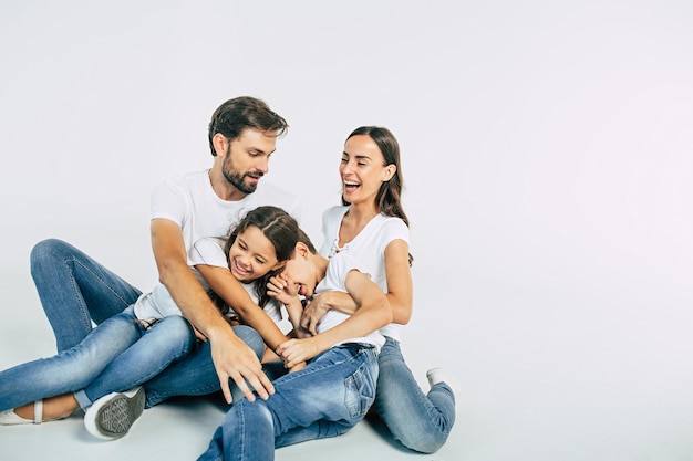 Hermosa y feliz familia joven sonriente con camisetas blancas se abrazan y se divierten juntos mientras están sentados en el suelo