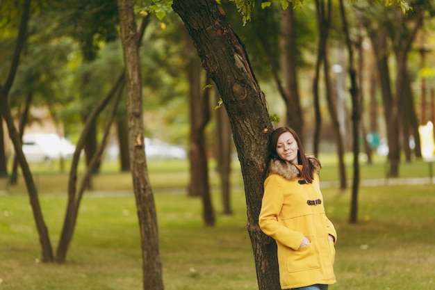 Hermosa feliz joven caucásica sonriente mujer de cabello castaño en abrigo amarillo, jeans, botas en bosque verde. Modelo femenino de moda con hojas doradas de otoño de pie y caminando en el parque a principios de otoño al aire libre.