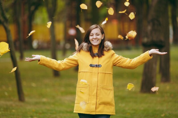 Hermosa feliz joven caucásica sonriente mujer de cabello castaño en abrigo amarillo, jeans, botas en bosque verde. Modelo femenino de moda vomitando hojas de otoño de pie y caminando en el parque a principios de otoño al aire libre.