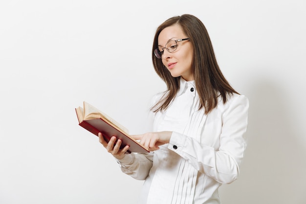 Hermosa feliz joven caucásica sonriente mujer de negocios de cabello castaño con camisa blanca y gafas leyendo libro rojo aislado en la pared blanca