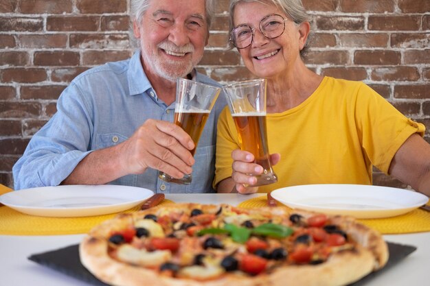 Hermosa feliz pareja de ancianos divirtiéndose juntos comiendo pizza Sentados en la mesa de casa brindando con un vaso de cerveza fondo de pared de ladrillo