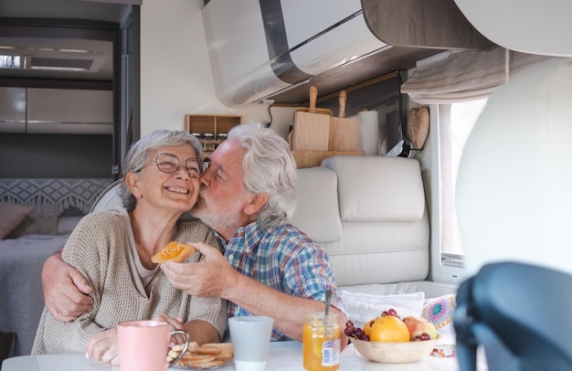 Hermosa feliz pareja caucásica senior en vacaciones de viaje sentado dentro de una autocaravana dinette disfrutando del desayuno juntos marido besa a su esposa