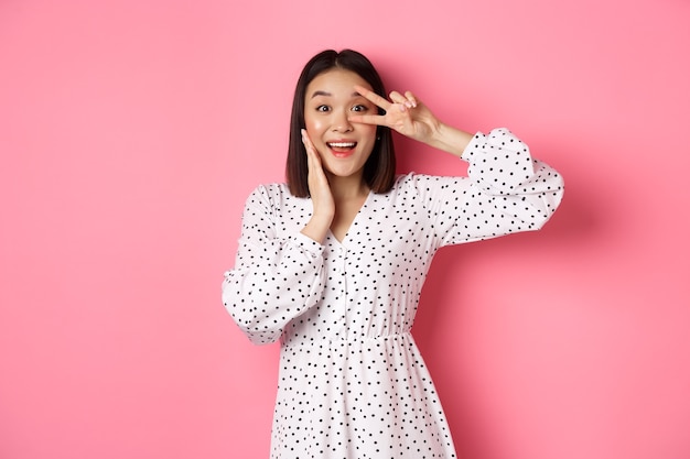 Hermosa y femenina mujer asiática mostrando el signo de kawaii en el ojo y mirando asombrado a la cámara, de pie en vestido sobre fondo rosa