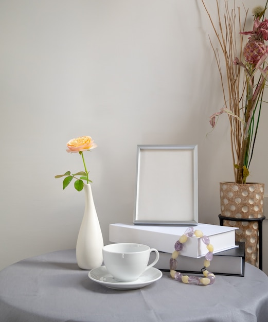 Hermosa flor color de rosa en florero blanco con libro de marco plateado y taza de café en la mesa de tela de lino