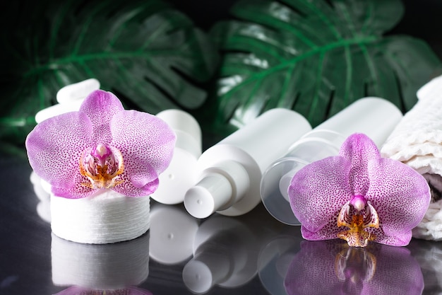 Hermosa flor de orquídea lila, botellas de cosméticos blancas, almohadillas de algodón con pila de piedras blancas y hojas de monstera sobre superficie negra.