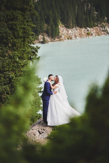 Hermosa foto de boda en el lago de la montaña. Feliz pareja asiática enamorada, la novia con vestido blanco y el novio en traje son fotografiados con el telón de fondo del paisaje kazajo