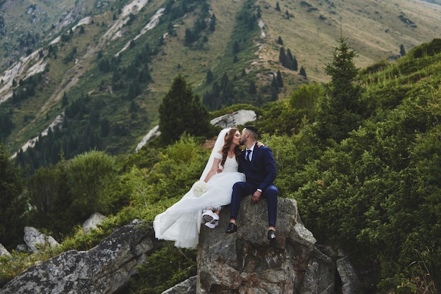 Hermosa foto de boda en el lago de la montaña. Feliz pareja asiática enamorada, la novia con vestido blanco y el novio en traje son fotografiados con el telón de fondo del paisaje kazajo