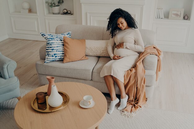 Hermosa foto dama afroamericana sentada en un sofá acogedor y esperando un bebé mirando hacia abajo en su campana