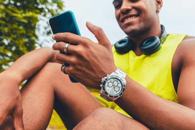 Hermosa foto de un hombre joven con auriculares sonriendo a su teléfono mientras está sentado en un parque