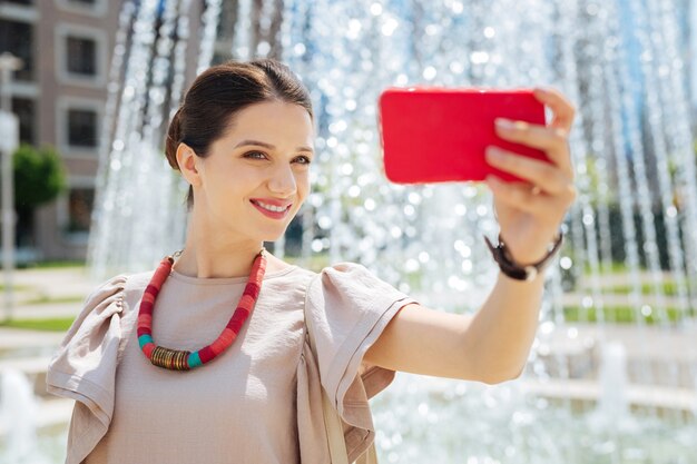 Hermosa foto. Mujer atractiva alegre tomando un selfie mientras está de pie cerca de la fuente