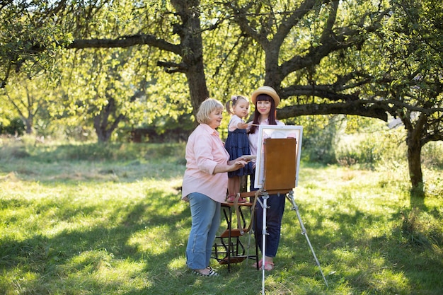 Hermosa foto de primavera de verano de familia feliz de tres generaciones, niña niño, madre joven y abuela madura, pasar tiempo al aire libre y pintar cuadros en caballete. Ocio familiar de verano.