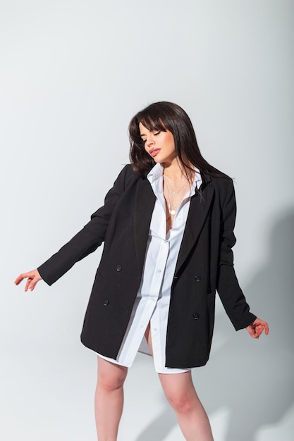 La hermosa y glamurosa chica de negocios de moda en un elegante blazer negro de gran tamaño y camisa blanca baila y posa sobre un fondo blanco en el estudio