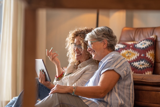 Hermosa hija adulta con madre anciana sentada en casa mirando redes sociales en tableta digital