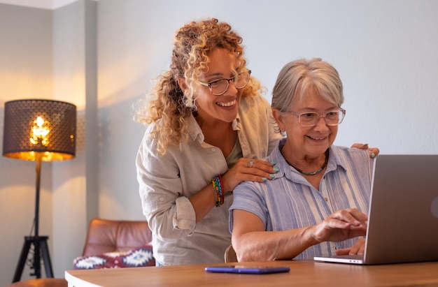 Hermosa hija adulta rubia enseñando a su madre cómo usar la red con una computadora portátil digital Concepto de compartir y complicidad