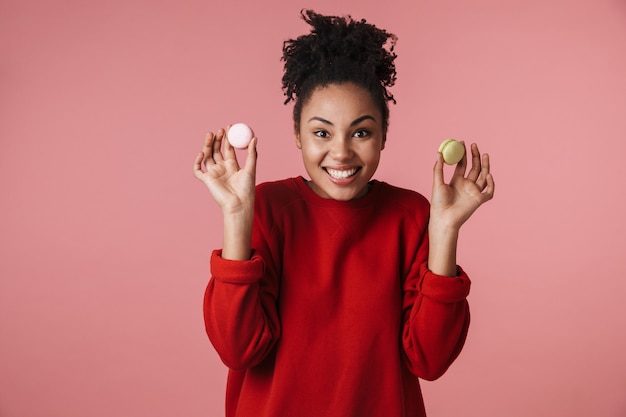 Hermosa increíble feliz emocionada joven africana posando aislada sobre pared rosa sosteniendo dulces macarrones.