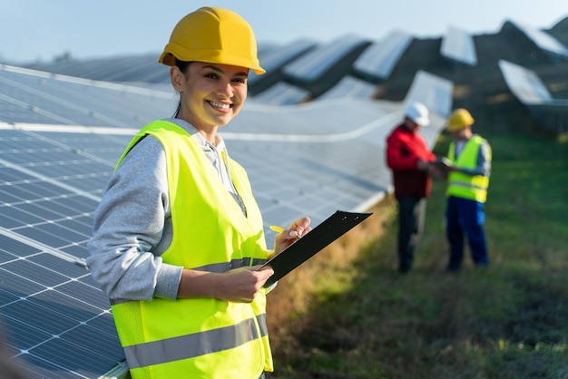 Hermosa ingeniera tecnóloga parada entre paneles solares y sosteniendo documentos Mujer con casco protector y uniforme sonriendo a la cámara