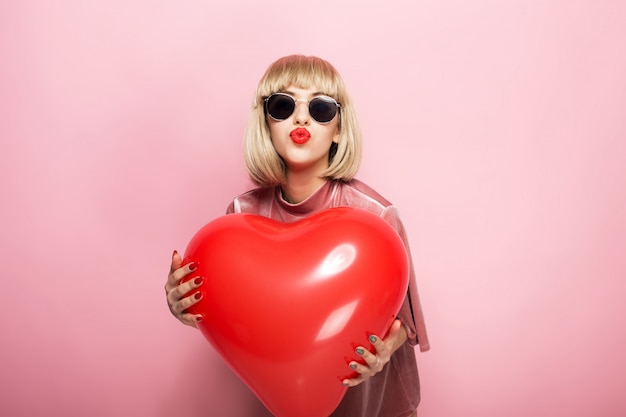 Hermosa joven abrazando en forma de corazón una bola roja y besos. Sobre un fondo rosa