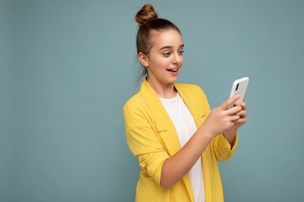 Hermosa joven adolescente vistiendo ropa casual que se encuentran aisladas sobre fondo navegando en Internet a través del teléfono mirando la pantalla del móvil.