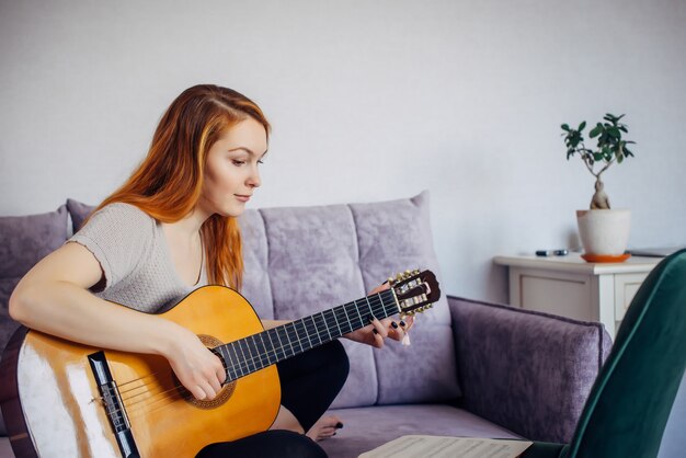 Hermosa joven adulta con el pelo largo tocando la guitarra, sentada en casa en el sofá, enfoque selectivo. Mujer seria toca las cuerdas, estudiando la melodía. Ocio en el hogar, pasatiempos, autodesarrollo.