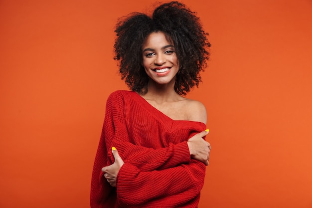 Hermosa joven africana alegre vistiendo suéter de pie islolated sobre pared roja, brazos cruzados