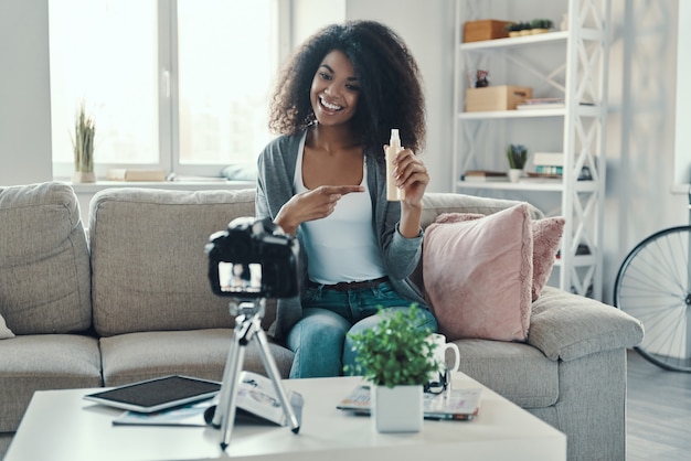 Hermosa joven africana apuntando a productos de belleza y sonriendo mientras hace videos de redes sociales