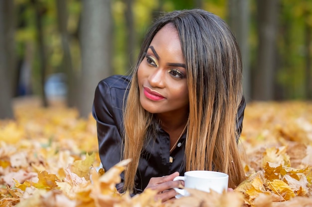 Hermosa joven africana bebiendo café de una taza blanca en el fondo de hojas de otoño en el parque