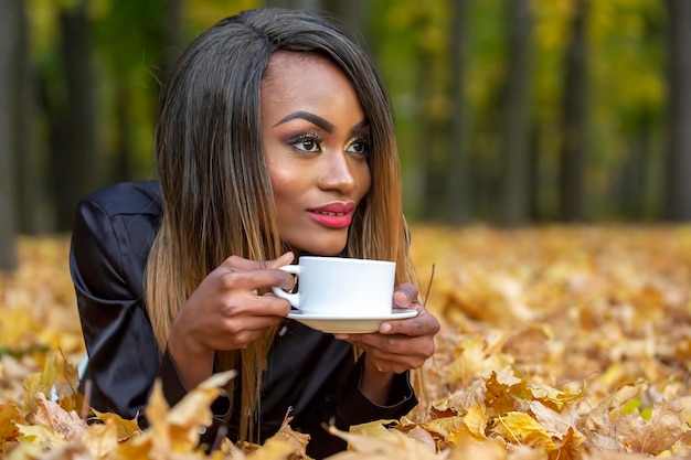 Hermosa joven africana bebiendo café de una taza blanca en el fondo de hojas de otoño en el parque