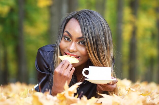 Hermosa joven africana comiendo galletas y bebiendo café de la taza blanca en hojas de otoño en el parque