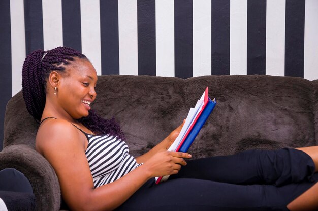 Hermosa joven africana sentada en un sofá en casa, emocionada por el libro que está leyendo.