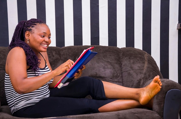 Hermosa joven africana sentada en un sofá en casa, emocionada por el libro que está leyendo.