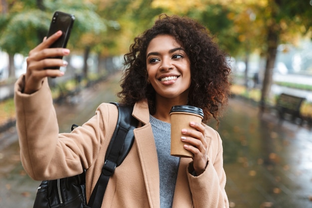 Hermosa joven africana vistiendo abrigo caminando al aire libre en el parque, llevando mochila, tomando un selfie