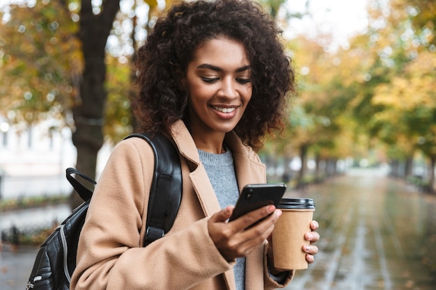 Hermosa joven africana vistiendo abrigo caminando al aire libre en el parque, llevando mochila, usando teléfono móvil, tomando café