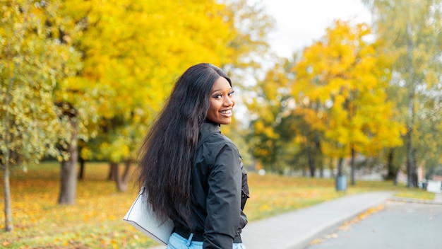 Hermosa joven afroamericana con una sonrisa en ropa de moda camina en un parque de otoño con increíble follaje amarillo