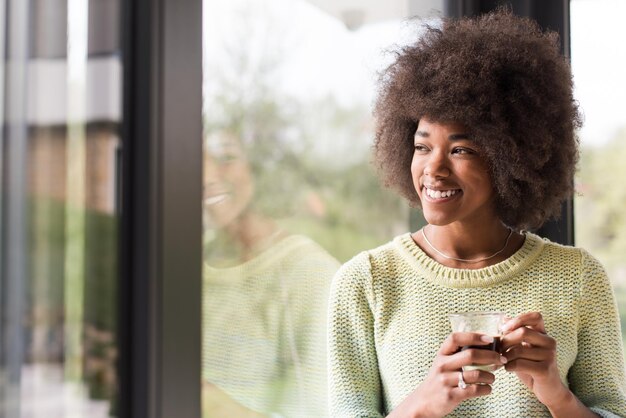 Hermosa joven afroamericana tomando café y mirando a través de una ventana en su casa de lujo